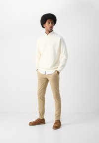 Cremefarvet sweatshirt med høj krave og diskret logo. Beige chinos og brune loafers fuldender outfittet. Modellen står på en hvid baggrund.