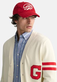Cappellino da baseball in corduroy rosso con il logo "Gant 1949", sovrapposto a una camicia a righe azzurre chiaro e a un cardigan crema con accenti rossi.