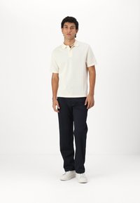 Cream-colored polo shirt with a textured pattern, short sleeves, and a collared neckline, paired with dark blue pants and white sneakers.