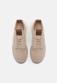 Beige textured sneakers with a knit upper, brown leather heel tab, flat laces, and a white sole with dark rubber accents.