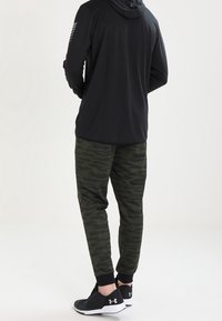 Mustat pitkät hihaisella hupparilla, jossa huppu ja sivu raidat, yhdistettynä tumman vihreisiin kamojogginghousuihin, lopputuloksena mustat urheilukengät.