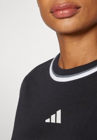 Μαύρο κοντομάνικο πουκάμισο με λευκή και γκρίζα ρίγα στον λαιμό, με λευκό λογότυπο που απεικονίζει τρεις ράβδους στο στήθος. Λείος υφασμάτινος ιστός.