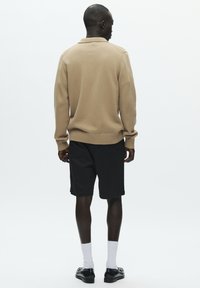 Pull tricot beige avec poignets et ourlet côtelés, associé à un short noir. Le mannequin porte des chaussettes blanches et des chaussures noires, vu de dos.