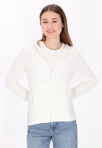Valkoinen vetoketjuhuppari, jossa on pehmeä pinta ja huppu, yhdistettynä vaaleaan paitaan ja sinisiin farkkuihin. Malli seisoo kädet lanteilla.
