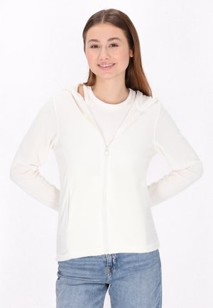 Weißer Reißverschluss-Hoodie mit weicher Textur und Kapuze, kombiniert mit einem hellen T-Shirt und blauen Jeans. Das Modell steht mit den Händen in den Hüften.