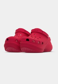 Crocs vermelhos brilhantes feitos de material leve, apresentando orifícios de ventilação, bico arredondado e uma tira com fivela no calcanhar para suporte.