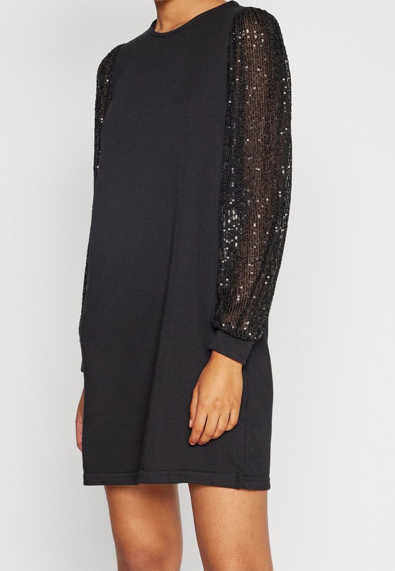 Femme portant une robe noire avec des manches longues transparentes et ornées de sequins, se tenant devant un fond clair uni.
