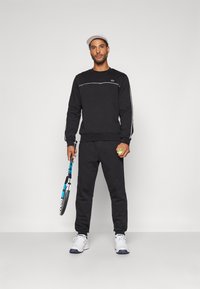 Svart hettegenser med hvit aksentstripe, kombinert med matchende svarte joggebukser. Iført en hvit caps, holder en tennisracket og ball.