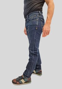 Jeans blu scuro con vestibilità slim, dotati di tasche frontali, leggere sfumature e dettagli di cucitura. Abbinati a scarpe sportive multicolori.