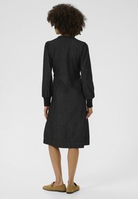 Zwarte spijkerdress met lange opgezette mouwen, een aansluitende taille, een rok tot de knie en manchetten. Opvallende stikdetails en een ontwerp van de kraag aan de achterkant.