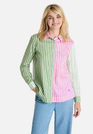 Frau lächelt, trägt ein Hemd mit grünen und pinken vertikalen Streifen sowie hellblaue Jeans und steht mit einer Hand in der Tasche.