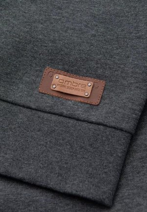Tissu gris avec une texture lisse, présentant une étiquette en cuir marron avec un texte en relief et des accents métalliques. Design simple et rectangulaire.