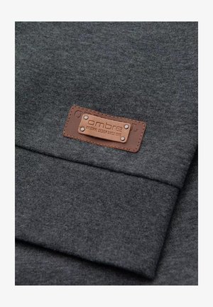 Tissu gris avec une texture lisse, présentant une étiquette en cuir marron avec un texte en relief et des accents métalliques. Design simple et rectangulaire.