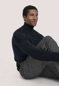 Schwarzer Rollkragenpullover aus weichem Stoff, kombiniert mit grauen strukturierten Hosen. Modell sitzt mit übergeschlagenen Beinen und schaut leicht nach links.