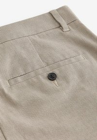 Beige teksturerede bukser med en baglomme, der har en sort fire-huls knap og en flad taljedetalje. Subtilt mønster tilføjer visuel interesse.