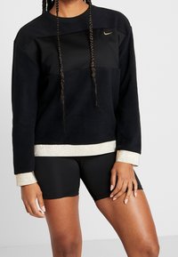 Musta fleece-huppari, jossa on kultaiset yksityiskohdat helman ja hihansuiden ympärillä; siinä on musta paneeli rinnassa ja brodeerattu logo vasemmassa yläkulmassa.