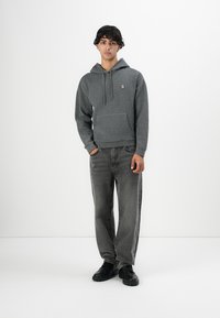 Young man standing, wearing a textured gray hoodie, loose dark gray jeans, and black boots against a plain white background.