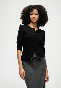 Črn dolg rokav kardigan s okroglim izrezom, gumbom za zapiranje in rebrastimi manšetami, v kombinaciji s sivo krilom na vrvico. Gladka tekstura.