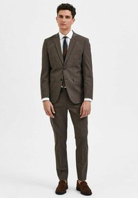 Bruin geruit pak met een slim fit, colbert met één knoop en bijpassende broek. Witte shirt en donkere stropdas met geometrisch patroon. Bruine loafers.