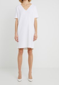 Robe blanche à manches courtes avec un décolleté en V profond, coupe ample et tissu lisse. Ourlet au genou, associée à des talons blancs à bout pointu.