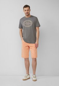 Graues Baumwoll-T-Shirt mit rundem Druckdesign. Pfirsichfarbene Jeansshorts. Lässige weiße Sneaker mit beigen Akzenten und Schnürsenkeln. Klarer Hintergrund.