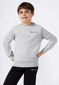 Jongen in grijze Champion-trui en zwarte Champion-broek staat met zijn handen op zijn heupen, glimlachend tegen een witte achtergrond.