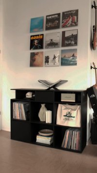 Czarna jednostka półkowa z ośmioma otwartymi przegródkami, zawierająca winyle, ceramiczny wazon, torbę materiałową oraz książkę z podpórką na górze.