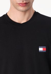 Svart bomulls t-skjorte med ribbet rund hals og en rektangulær logo-patch i rødt, hvitt og blått på venstre bryst.