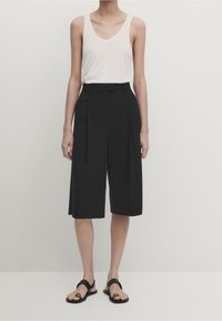 Mustat culotte-housut laskostetulla designilla, yhdistettynä valkoiseen hihattomaan toppiin. Kangas näyttää pehmeältä ja kevyeltä. Mustat sandaalit täydentävät asun.