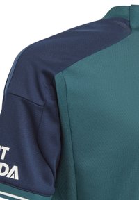 Urheilu t-paita lyhyillä hihoilla, jossa on teksturoitu tumman teal-värinen vartalo ja laivastonsininen hartialäppä, sekä valkoinen logotyyppi edessä.