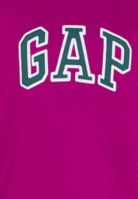 Sweat-shirt fuchsia avec le mot "GAP" en grandes lettres vertes brodées, bordées de blanc. Tissu doux avec une texture lisse.