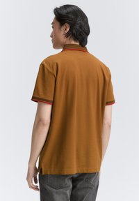 Polo de manga corta marrón con un cuello y mangas cortas a rayas rojas en contraste. Está hecho de una tela suave y ligera.