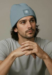 Gray ribbed knit beanie with a square patch displaying a compass logo. Worn with a gray t-shirt, featuring a smooth texture and a fitted shape.