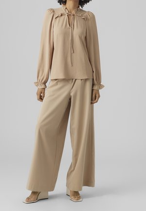 Blouse beige à col volanté avec détail à nouer, associée à un pantalon beige à jambes larges. Texture de tissu lisse avec une coupe décontractée. Détails clairs sur les manches.