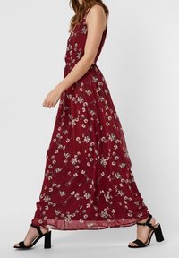 Femme marchant dans une robe maxi sans manches bordeaux à motif floral blanc et des sandales à talons noirs, vue de côté.