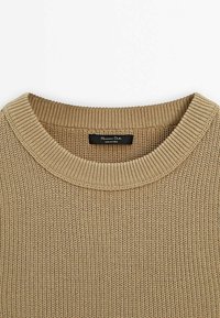 Beige neulepusero, jossa on ribattu pyöreä kaula-aukko. Ominaisuuksia ovat löysä neulostekstuuri ja musta etiketti, jossa lukee "Massimo Dutti, Valmistettu Kiinassa."