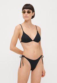 Black ribbed bikini with triangular top and adjustable straps; bottoms feature tie sides. Model wearing oval sunglasses.