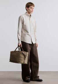 Μπεζ πουκάμισο με κουμπιά, καφέ φαρδιές παντελόνες, κρατώντας μια μαλακή τσάντα ταμπά με καφέ λεπτομέρειες και φερμουάρ. Λιτή σχεδίαση με λείο ύφασμα.