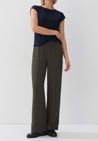 Osoba oblečená v námořnickém tričku s krátkým rukávem, olivově zelených širokých kalhotách a černých mokasínech na jednolitém světlém pozadí.