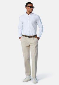 Camicia bianca con bottoni, pantaloni beige, cintura marrone e scarpe da ginnastica bianche. Il modello è in piedi con le mani nelle tasche, indossa occhiali da sole scuri.