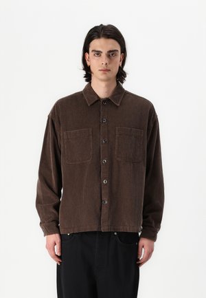 Camisa de pana marrón con cuello tipo polo, cierre de botones y dos bolsillos frontales. Presenta una textura de rayas verticales y un corte relajado.