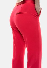 Pantaloni rossi su misura con una texture liscia, dotati di una cintura in satin e una tasca posteriore. Il design è aderente in vita, svasandosi leggermente alle gambe.