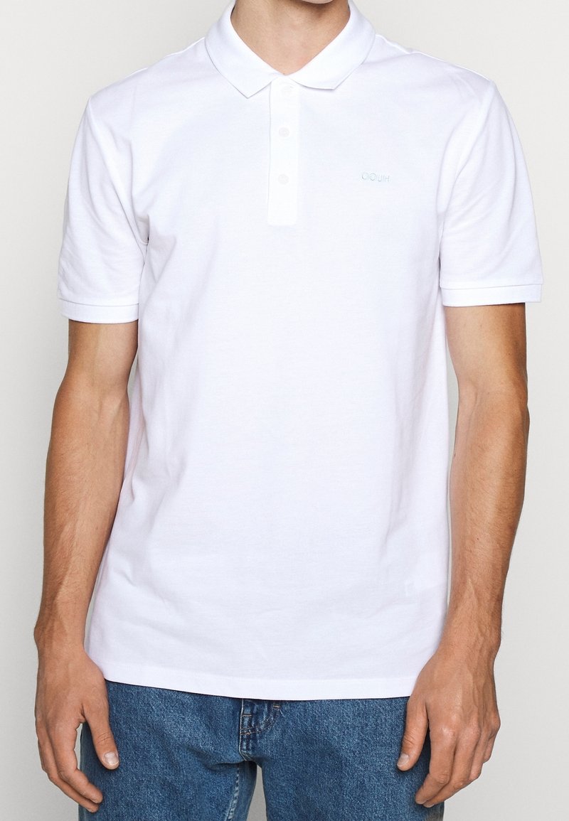 Homme portant un polo blanc uni à manches courtes et un jean bleu, debout devant un fond uni.