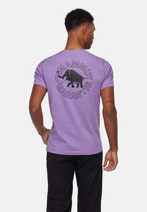 Lila T-Shirt mit kurzen Ärmeln, das ein schwarzes Mammut-Graphic und den Text "GARANTIE" auf dem Rücken zeigt. Aus weichem Baumwollstoff gefertigt.