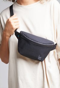 Borsa a tracolla nera in tessuto con chiusura zip e forma piatta. Presenta un logo e accenti colorati sulla tracolla. Indossata a tracolla.