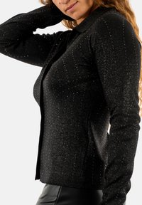 Cardigan nero a maglia con colletto a punta, caratterizzato da una texture scintillante e da punti argentati sparsi su tutto il tessuto. Stile aderente.