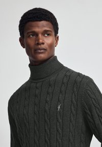 Dunkelgrauer, kabelstrickartiger Rollkragenpullover mit geripptem Kragen, der ein kleines Logo-Detail auf der Brust und ein strukturiertes Muster über das gesamte Kleidungsstück aufweist.