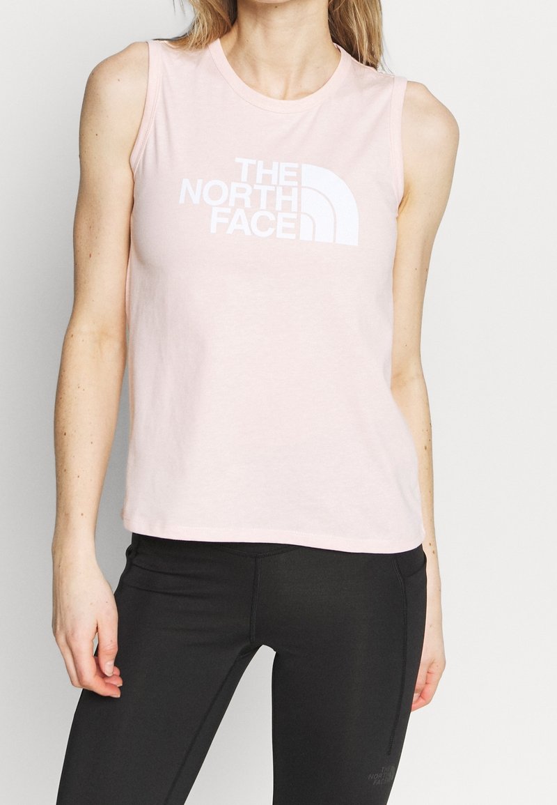 Femme portant un débardeur rose clair avec le logo The North Face et des leggings noirs sur un fond uni.