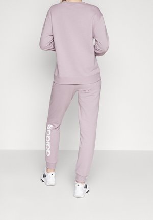 Personne portant un pantalon de survêtement Adidas violet clair et un sweat-shirt assorti, debout de dos, avec des baskets blanches Adidas.