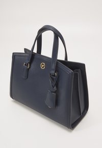 Sac à main en cuir texturé bleu marine avec double poignées, emblème circulaire doré et étiquette en cuir suspendue sur un fond neutre.